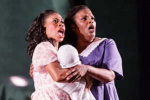Chrystal E. Williams as Rebecca Parker in Opera Philadelphia's 2015 premiere performance of Charlie Parker's Yardbird. Photo by Dominic M. Mercier. Williams is pictured here in a pink and white floral dress, holding a baby. Angela Brown as Charlie Parker's mother, is pictured in a purple print dress, holding both Williams and the baby. They sing, fiercely, both with mouths open in determination and perseverance.