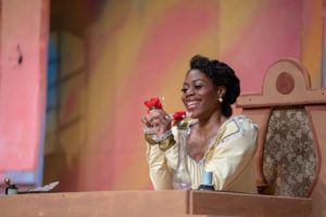 Chrystal E. Williams as Rosina in Northern Lights Music Festival's 2018 production of Il barbiere di Siviglia. She is pictured here in a pale yellow gown fringed in lace, face lit up in a smile as she stares at a red flower while seated at a desk. Photo by Aaron Fagerstrom.