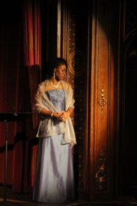 Chrystal E. Williams as Charlotte in the 2017 joint concert production of Werther by Baltimore Concert Opera and Opera Delaware. Photo by Courtney Kalbacker. Williams is pictured here in a periwinkle gown, dangling diamond earrings, ruby ring, diamond bracelet, and shimmery silver shawl. Her face is pensive as it looks downward. She is backed against a dark brown facade.