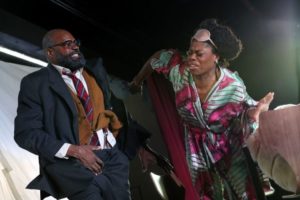 Chrystal E. Williams as Lady Macbeth with Eric Greene as Boris. Lady Macbeth rips her hand away from Boris as he tries to put it down the front of his pants. She is seen with a pink eye mask and a foam green and pink silk robe covered in mushrooms. Boris is seen in a suit with overcoat, glasses, and tie. Photo by Adam Fradgley